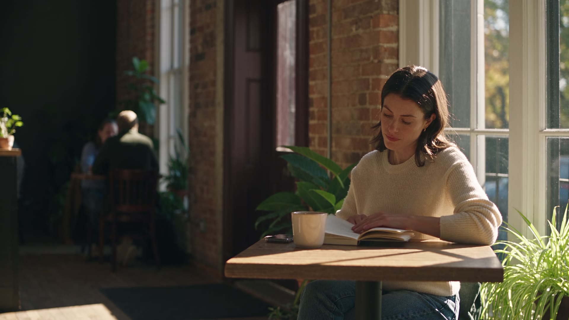 A woman in a cream knit reads with coffee at a sunny café window table